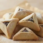 צילום קולינרי מקצועי של ערימת אוזני המן זהובות במילוי פרג או שוקולד, זרויות באבקת סוכר ומונחות על נייר אפייה על רקע בהיר ורך.