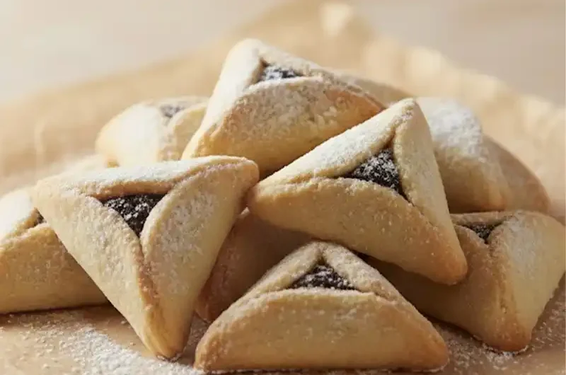 צילום קולינרי מקצועי של ערימת אוזני המן זהובות במילוי פרג או שוקולד, זרויות באבקת סוכר ומונחות על נייר אפייה על רקע בהיר ורך.