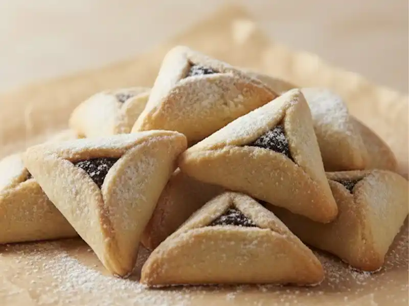 צילום קולינרי מקצועי של ערימת אוזני המן זהובות במילוי פרג או שוקולד, זרויות באבקת סוכר ומונחות על נייר אפייה על רקע בהיר ורך.