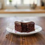 פרוסת עוגת שוקולד עסיסית מצופה בשכבה נדיבה של גנאש שוקולד פרווה מבריק, מונחת על צלחת לבנה על גבי שולחן עץ.